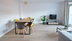 A modern living room with a wooden dining table, yellow chairs, a grey sofa, a TV on a wooden stand, and a potted plant.