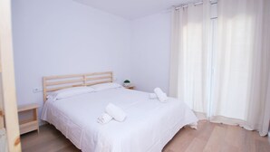 A neatly made bed with white linens and two rolled towels, a bedside table with a small plant, and a window with white curtains.