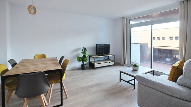 A modern living room with a wooden dining table, yellow chairs, a grey sofa, a television on a low cabinet, and a large sliding glass door to the outside.