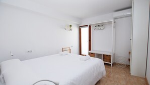 Una habitación de hotel limpia y blanca con una cama grande, un armario y una pequeña estantería con plantas.