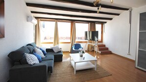Una sala de estar con sofá, mesa de centro y televisor. Se observan ventanas con cortinas, una escalera y techo de madera.