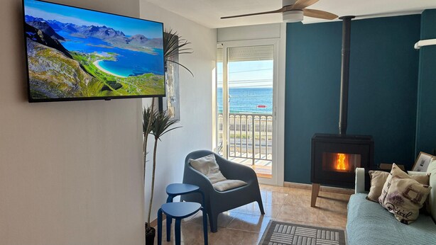 Una sala de estar con televisor mostrando un paisaje panorámico, chimenea, sofá, sillón y balcón con vista al mar.