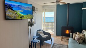 A living room with a TV displaying a scenic landscape, a fireplace, a sofa, a chair, and a balcony with a view of the sea.