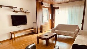 Una sala de estar con piso de madera, un sofá, una mesa de centro, un banco, un televisor y una ventana con cortinas.