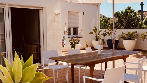 Un patio al aire libre con una mesa de madera y sillas blancas, rodeado de plantas en macetas y un paraguas blanco.