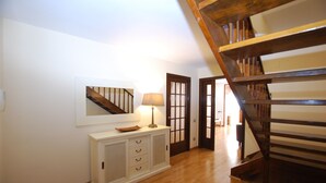 Un pasillo con piso de madera, un mueble aparador blanco, un espejo y una lámpara. Se ve una escalera de madera al fondo.