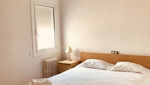 Un dormitorio con cama, ventilador de techo, mesita de noche con lámpara y ventana con persianas.