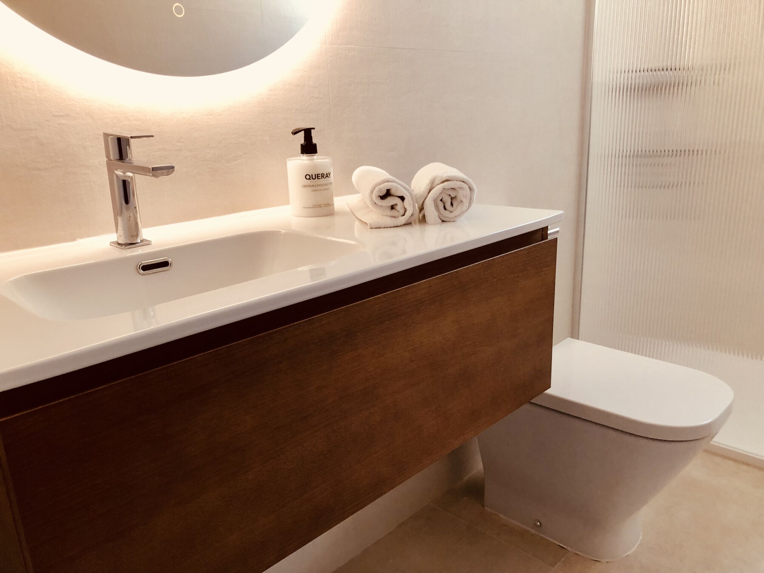 A modern bathroom with a white sink, a mirror, a bottle of hand soap, and two rolled towels.