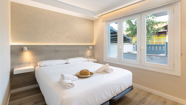 A modern hotel room with a large bed, a wooden headboard, a bedside table, and a window with a view of the outside. - Mestral - Aparthotel with Swimming Pool (Vilassar de Mar)