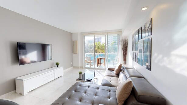 Ein modernes Wohnzimmer mit Fernseher, Sofa, Couchtisch und einem Balkon mit Blick auf Palmen und Gebäude.