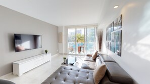 A modern living room with a flat-screen TV, a sofa, a coffee table, and a balcony with a view of palm trees and buildings.