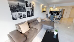 Ein modernes Wohnzimmer mit einer Eckcouch, einem Essbereich mit einem Glastisch und einer Küche im Hintergrund.