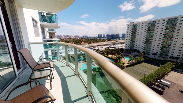 A balcony with a glass railing overlooking a swimming pool and a tennis court, with high-rise buildings in the background.