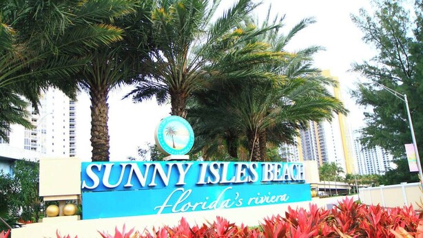 Señal de Sunny Isles Beach, Florida, Riviera, con palmeras y follaje rojo en primer plano.