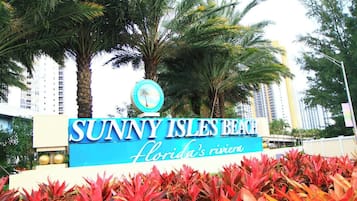 A sign for Sunny Isles Beach, Florida's Riviera, with palm trees and red foliage in the foreground.