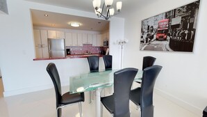 Una cocina moderna con armarios blancos, un refrigerador de acero inoxidable y una encimera roja. Hay una zona de comedor con una mesa de vidrio y sillas negras. En la pared, se muestra una escena callejera en blanco y negro.