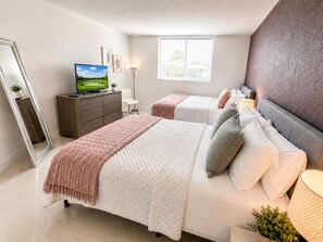 A bedroom with two beds, a TV, a dresser, and a window with blinds.