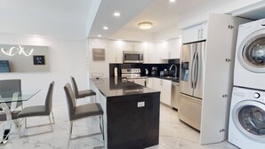 A modern kitchen with a central island, stainless steel appliances, and a dining area with glass tables and chairs. - OR1019 - Modern Ocean View Apt  Steps to the Beach (Sunny Isles)