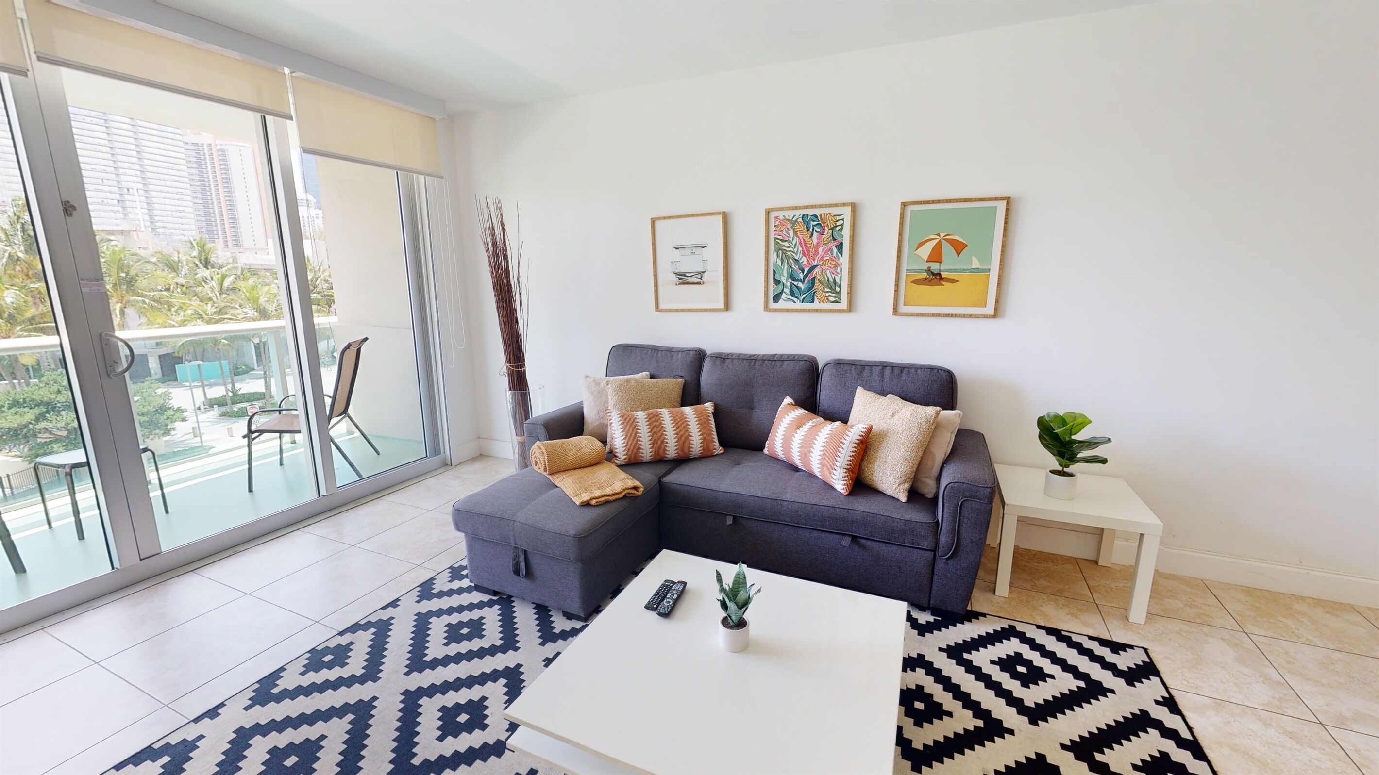 A modern living room with a grey sectional sofa, patterned rugs, and a balcony with a view of palm trees and buildings.