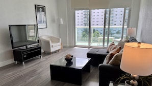 Una sala de estar moderna con una televisión de pantalla plana, un sillón blanco, un sofá negro, una mesa de centro de vidrio y un ventanal con vistas a un rascacielos.