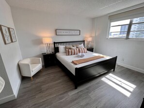 A modern bedroom with a black bed, white walls, and a large window.