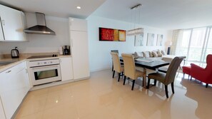 A modern kitchen with white cabinets, a stainless steel range hood, and a built-in oven. There is a dining area with a table and chairs, and a red chair in the corner. - Ocean Reserve 620 - Modern  Bay View Apartment! (Sunny Isles)