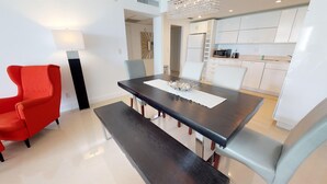 Un comedor moderno con una mesa de madera oscura, sillas blancas y un sillón rojo. Un candelabro cuelga encima, y se ve una cocina con armarios blancos al fondo.