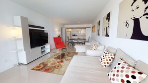Un salón moderno con un televisor de pantalla plana, una silla roja, un sofá blanco con cojines estampados y un comedor al fondo.