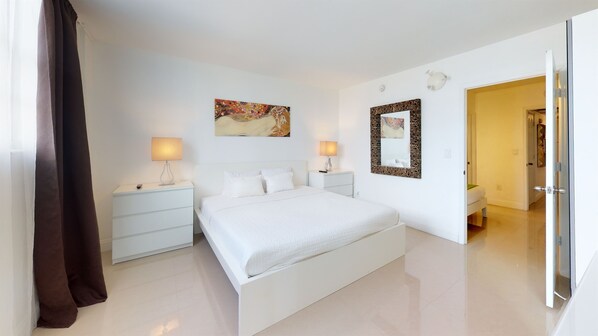 A modern bedroom with a large bed, two bedside tables with lamps, a decorative mirror, and a painting on the wall. - Ocean Reserve 620 - Modern  Bay View Apartment! (Sunny Isles)