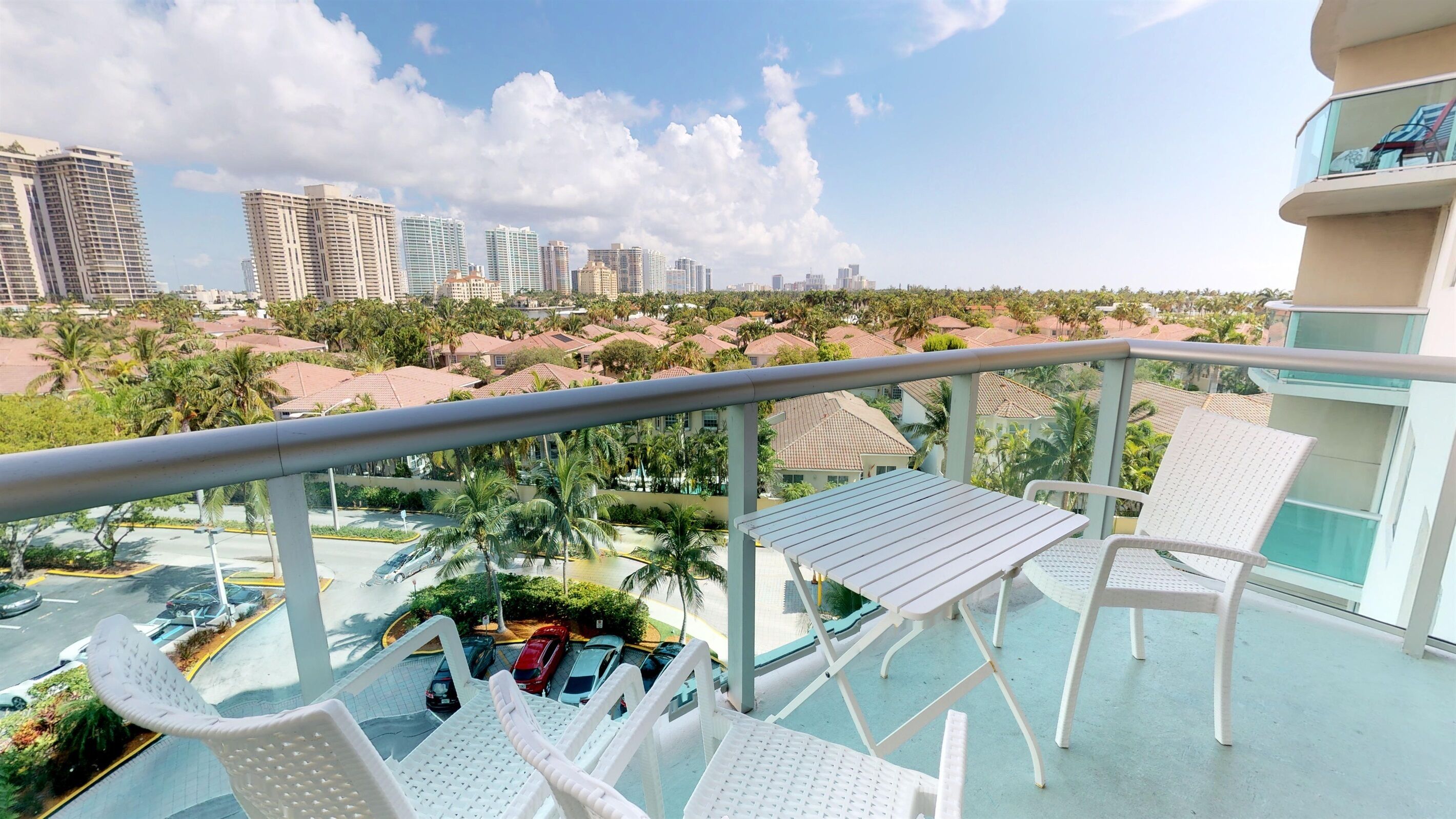 Ein Balkon mit weißen Stühlen und einem Tisch, mit Blick auf einen Parkplatz und eine Skyline mit Palmen und Hochhäusern.