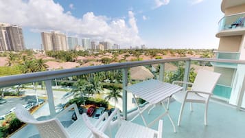 Ein Balkon mit weißen Stühlen und einem Tisch, mit Blick auf einen Parkplatz und eine Skyline mit Palmen und Hochhäusern.