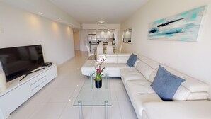 A modern living room with a flat-screen TV, a glass coffee table, a white sofa, and a kitchen in the background. - One Bedroom Ocean View  OR-925 (Sunny Isles)