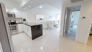 A modern kitchen with stainless steel appliances, a central island, and a dining area with white furniture. - One Bedroom Ocean View  OR-925 (Sunny Isles)