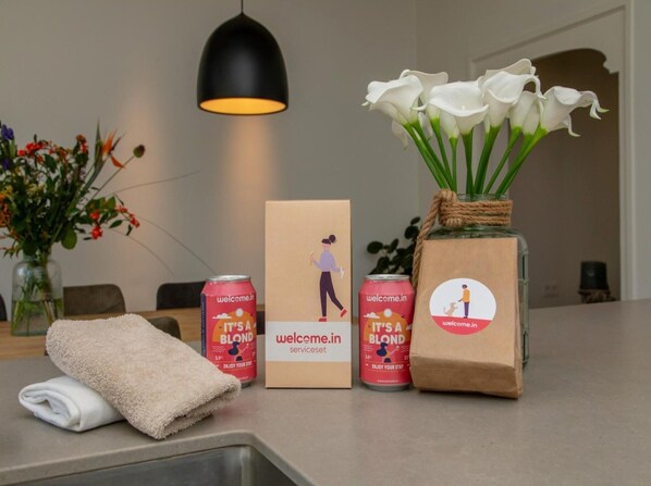 Ein Hotel-Empfangsbereich mit einer schwarzen Pendelleuchte, einer Vase mit weißen Blumen, einem zusammengerollten Handtuch und zwei Dosen des Getränks 'It's a Blond'.