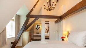 Un dormitorio con una cama grande, una araña, un viguelo de madera y vistas a un baño con espejo y lavamanos.
