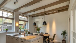 Una cocina moderna con una isla central, armarios de madera, una zona de comedor con mesa y sillas, iluminación colgante y vistas al exterior a través de amplios ventanales.
