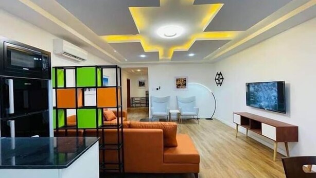A modern living room with a sofa, a TV, and a kitchen area with a fridge and a coffee machine. - Noktel Resort Apartment  (Abuja)