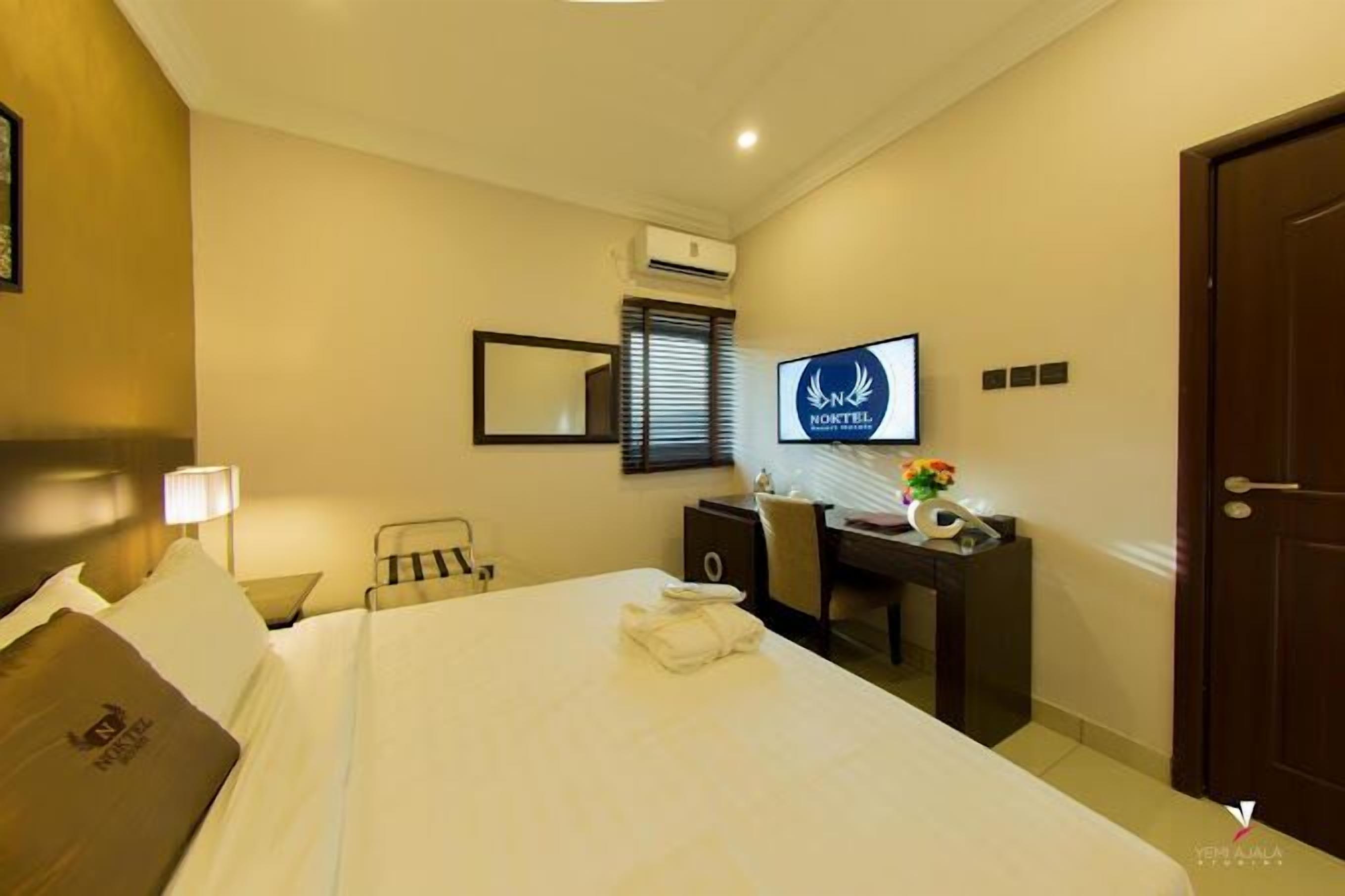 A hotel room with a bed, a desk, a chair, a TV, and an air conditioning unit.