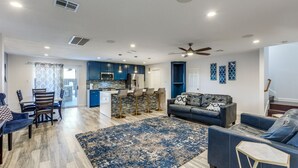 A modern living room with a blue sofa, a dining area with a wooden table and chairs, a kitchen with blue cabinets, and a staircase with a blue accent wall. - Spacious Home with Gorgeous Backyard Kitchen, Close to Top Shopping Destinations (Leander)