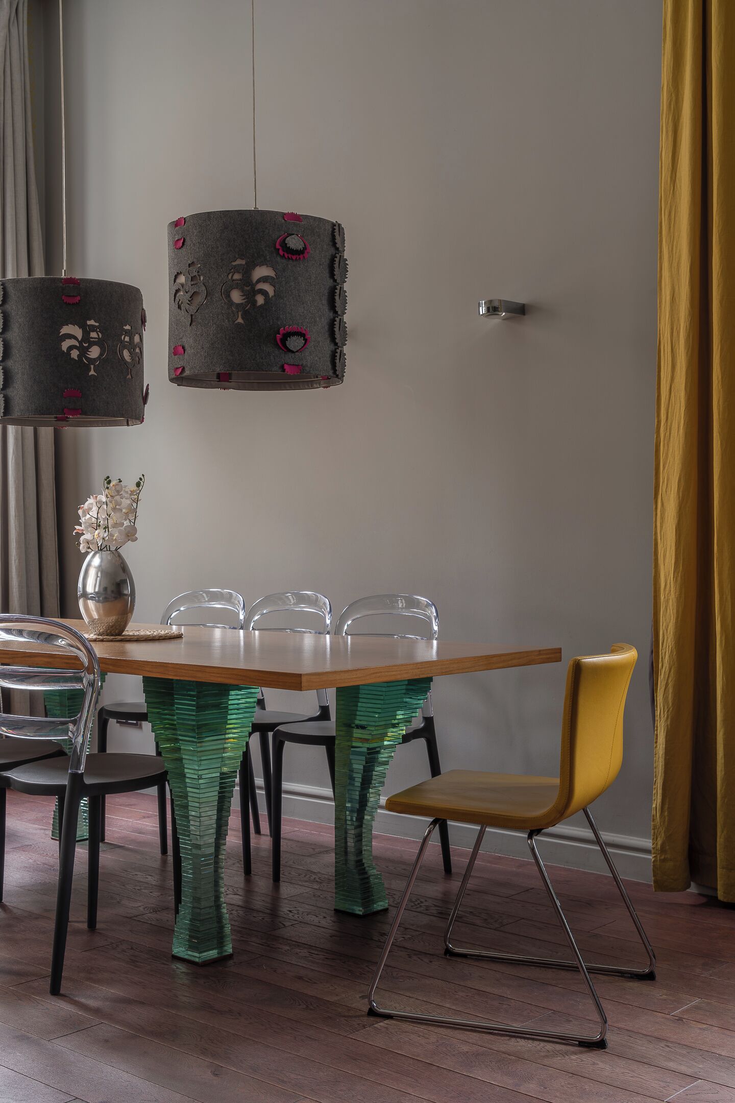 A dining area with a wooden table, clear chairs, and a yellow chair. There are two hanging lamps with patterned shades.