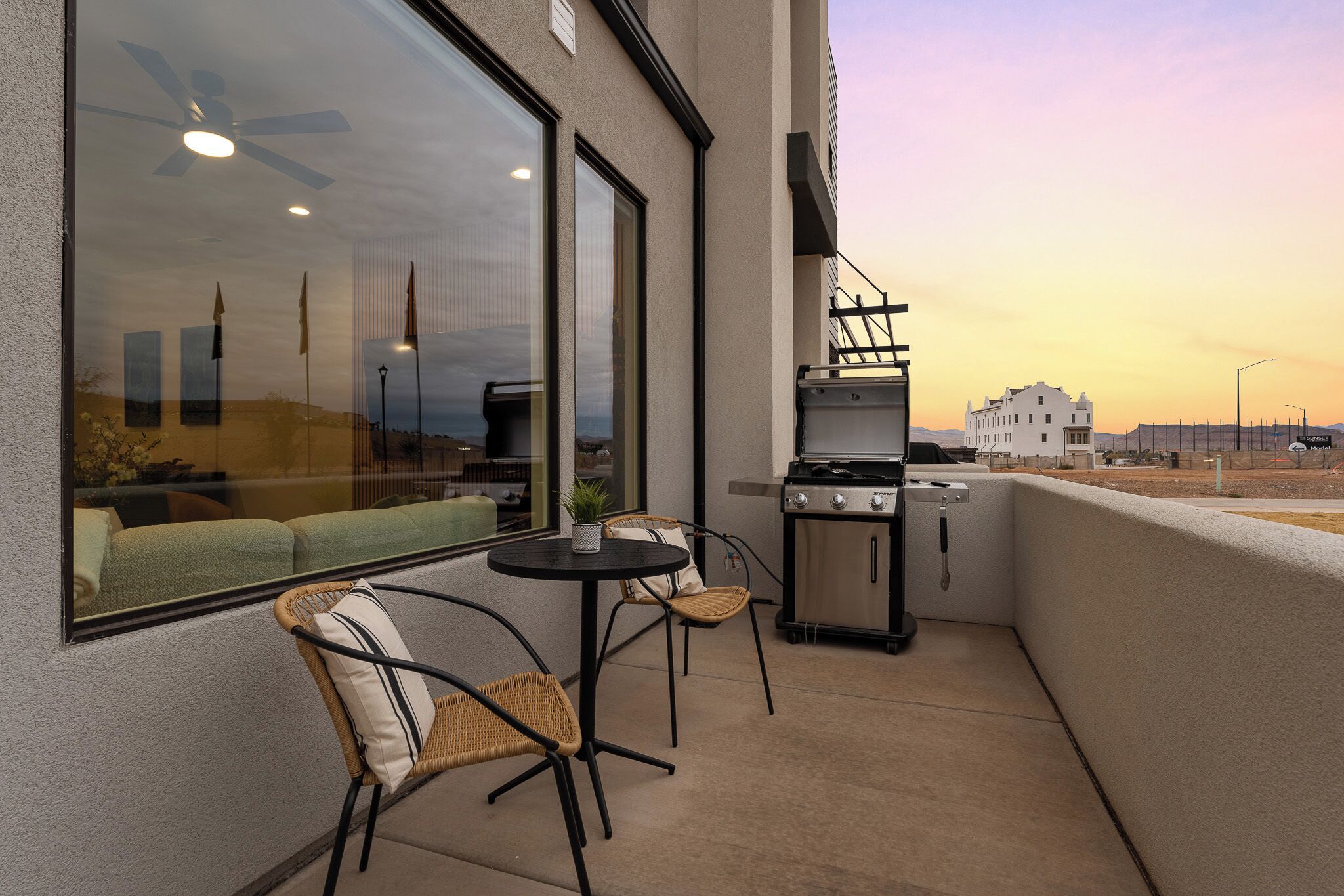 A balcony with a grill, wicker chairs, a small table, and a large window overlooking a desert landscape.