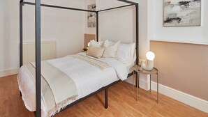 Een hemelbed met een witte spreideken en kussens, een nachtkastje met een lamp, en een radiator op de achtergrond.