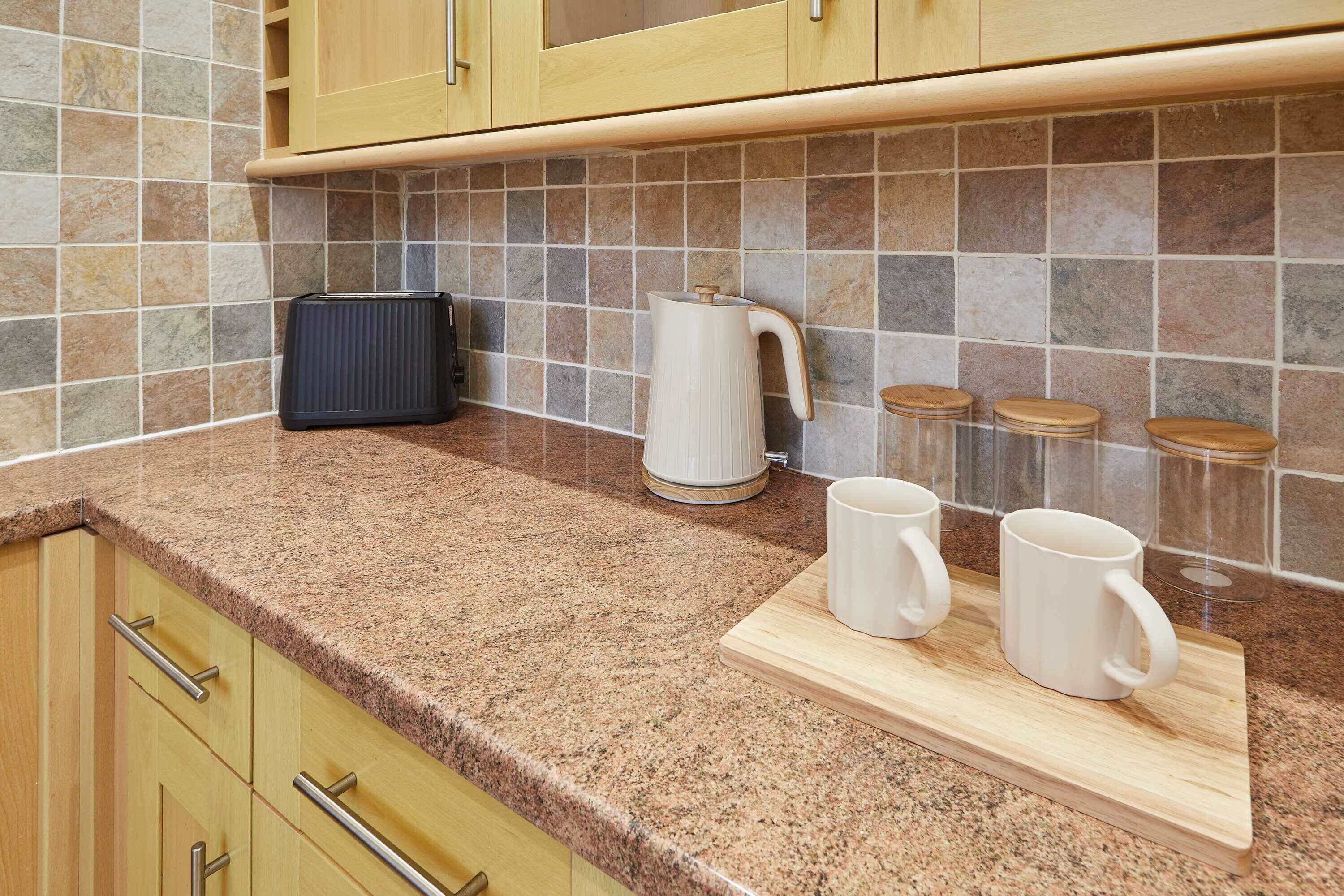Una cocina con encimeras de granito, armarios de madera, una cafetera y una tabla de cortar con tazas.