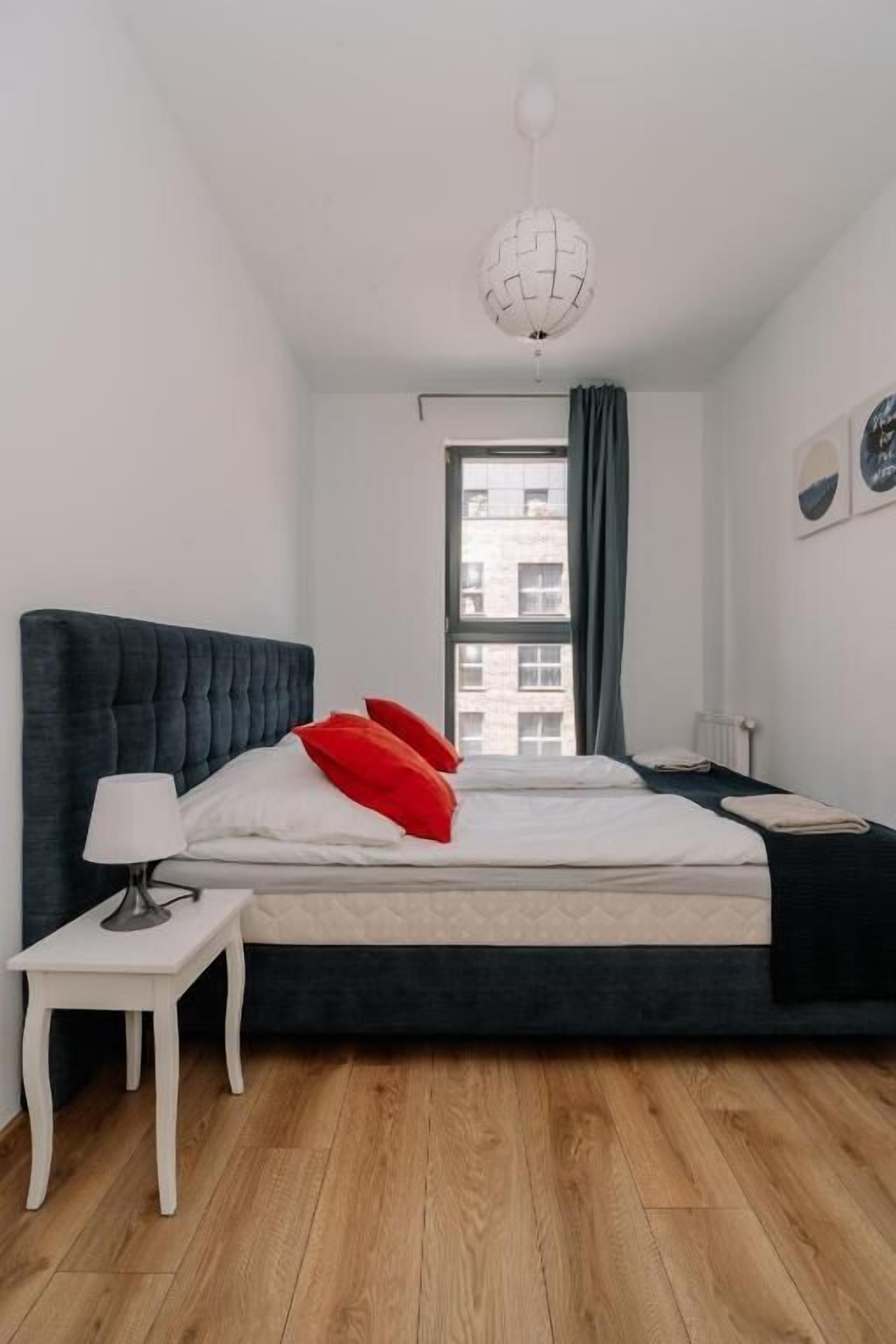 Una camera da letto con un letto grande, un comodino con una lampada, una finestra con tende e vista sui palazzi.
