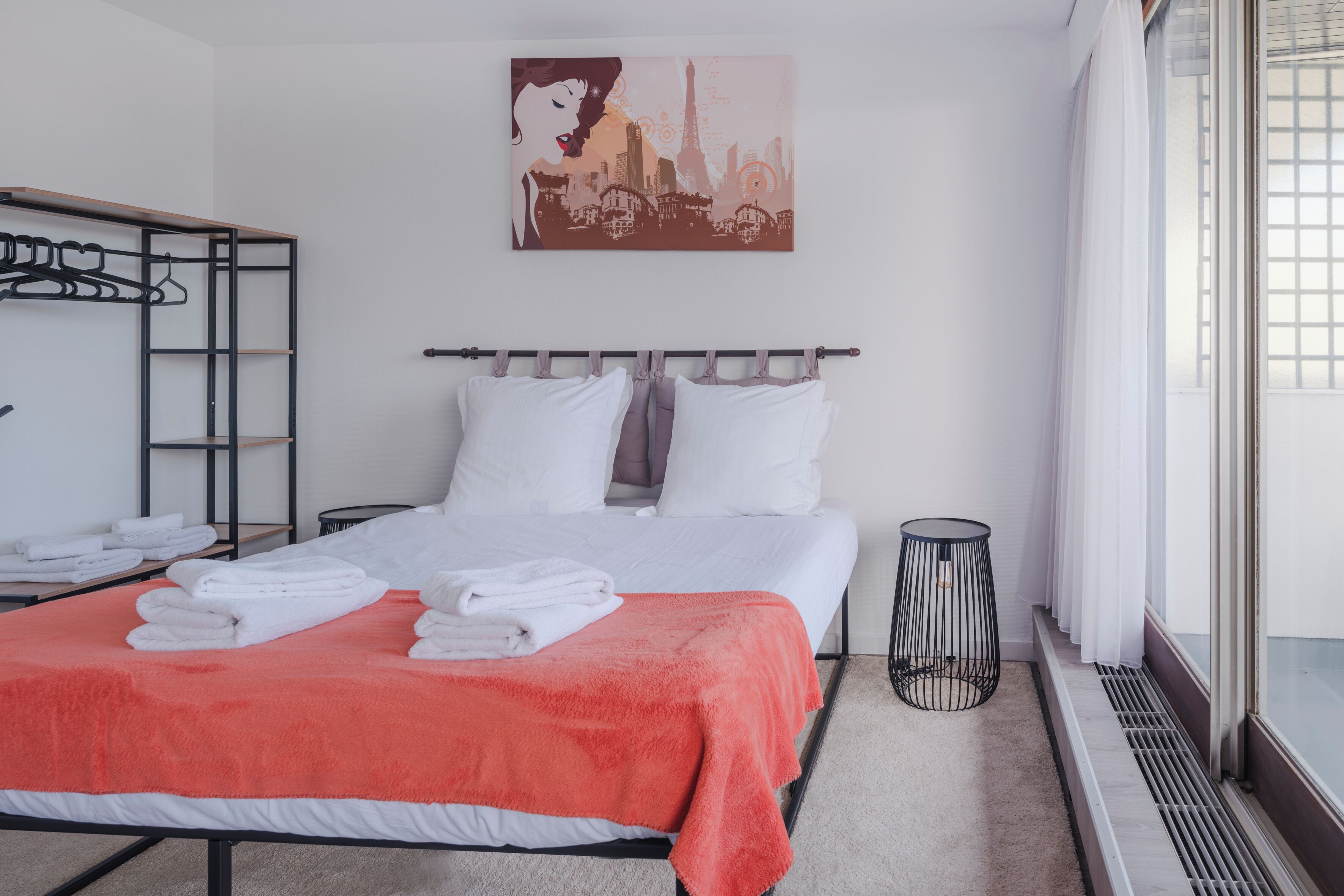 Ein Hotelzimmer mit Bett, weißer Bettwäsche, einer roten Decke, einem Stadtbildgemälde, einem Kleiderständer und einem Fenster mit Jalousien.