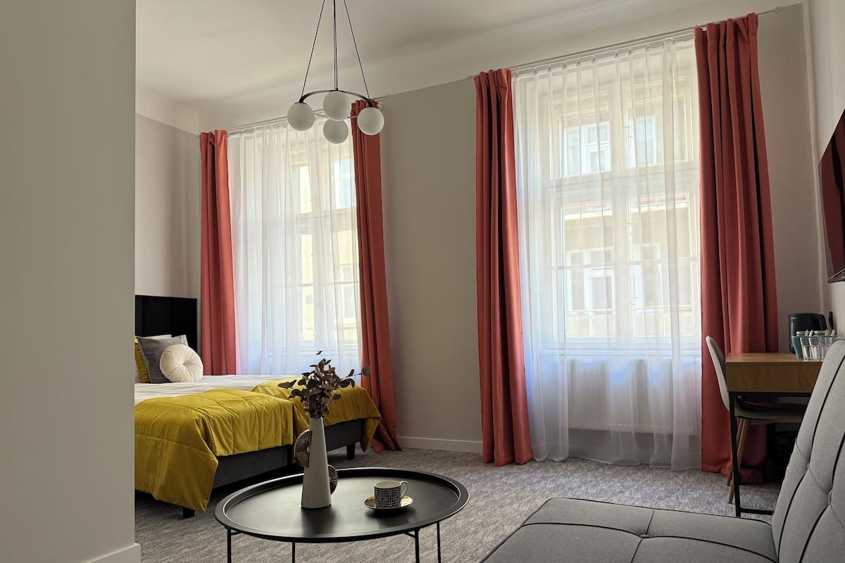 Une chambre d’hôtel moderne, équipée d’un lit, d’une table ronde agrémentée d’un vase et d’une tasse, d’un canapé et de grandes fenêtres avec des rideaux légers.