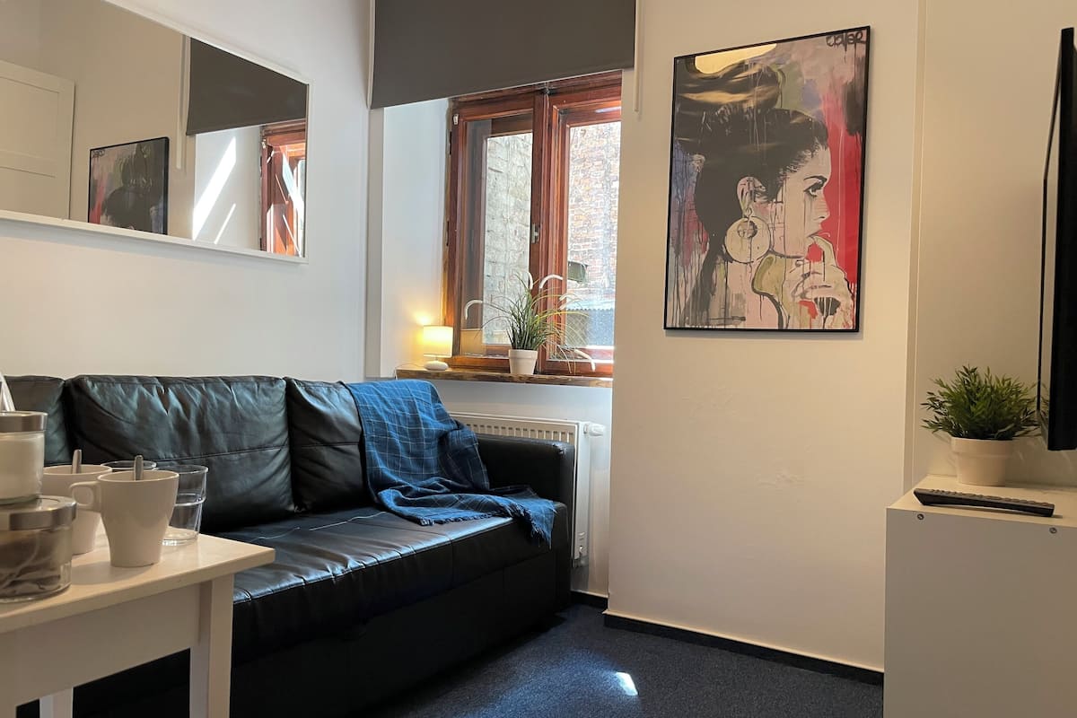 Un salon moderne avec un canapé en cuir noir, une table basse blanche et une grande fenêtre donnant sur un immeuble en briques.