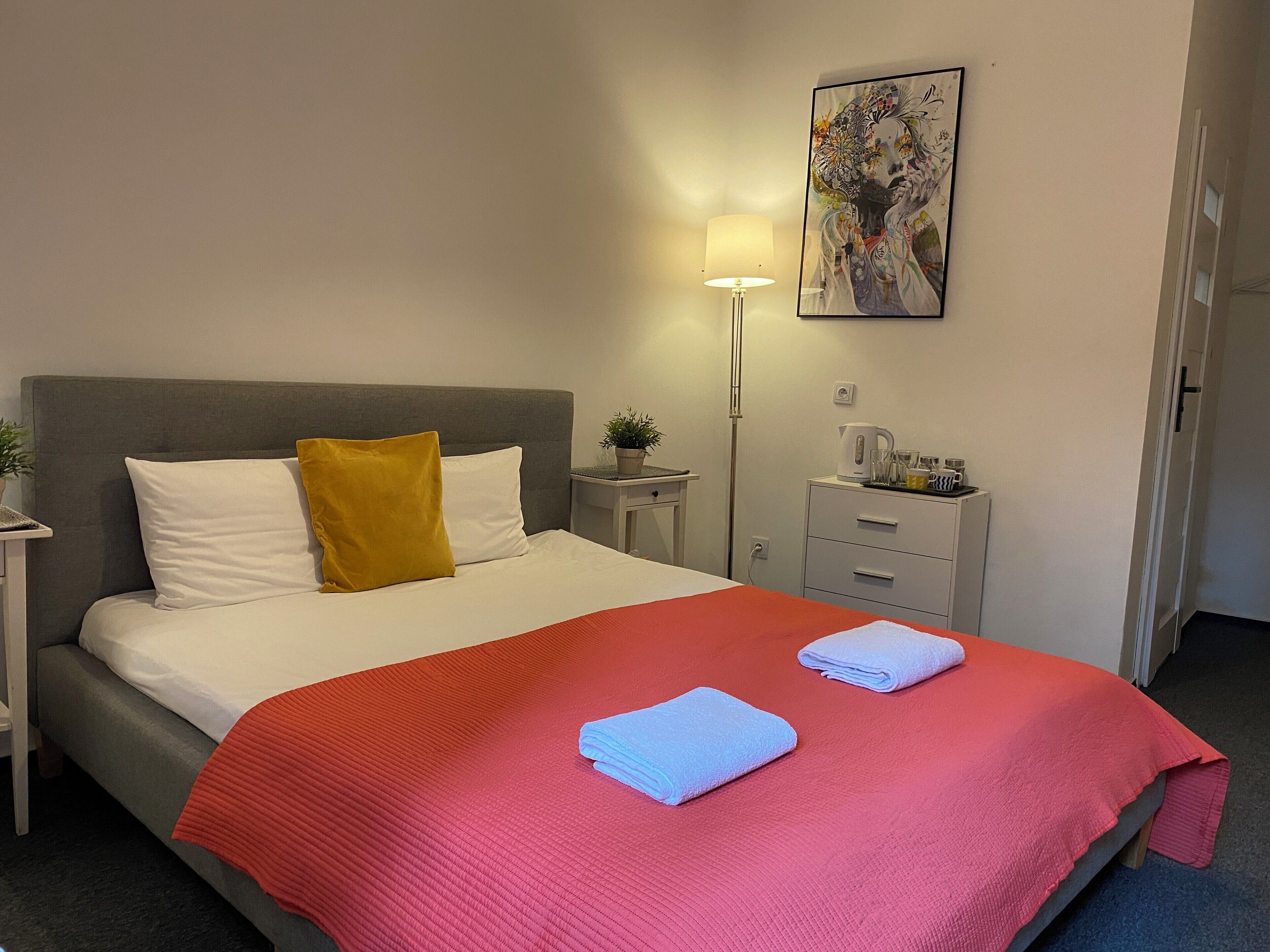 Una cama bien hecha con una manta roja, almohadas blancas y un cojín decorativo amarillo. Hay una mesita de noche pequeña con una lámpara y una planta en maceta. Un cuadro cuelga en la pared sobre la cama.