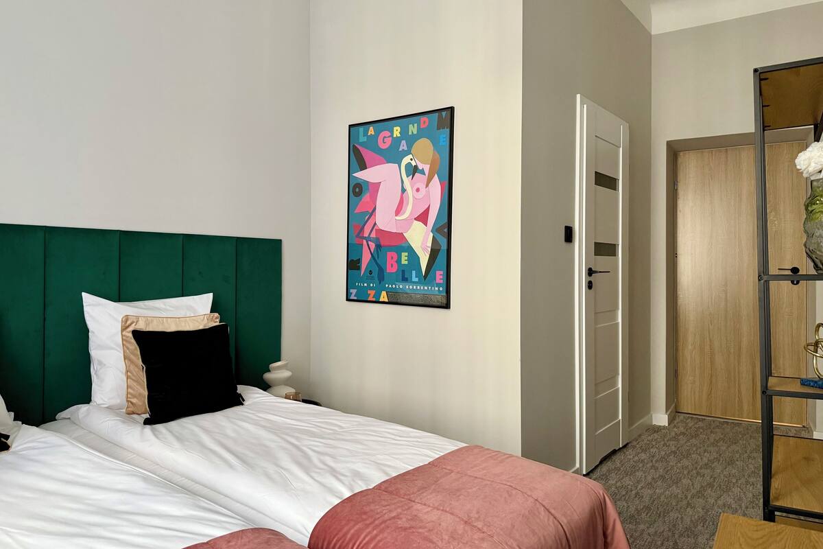 Une chambre d’hôtel avec un lit, une tête de lit verte, une affiche au mur et une porte en bois.