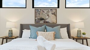 A neatly made bed with a white blanket and a rolled towel, flanked by two nightstands with lamps and a small plant. A large abstract painting hangs above the bed, and the room has a clean, modern aesthetic. - 2605 E 4th xxxxxxxxx (Austin)